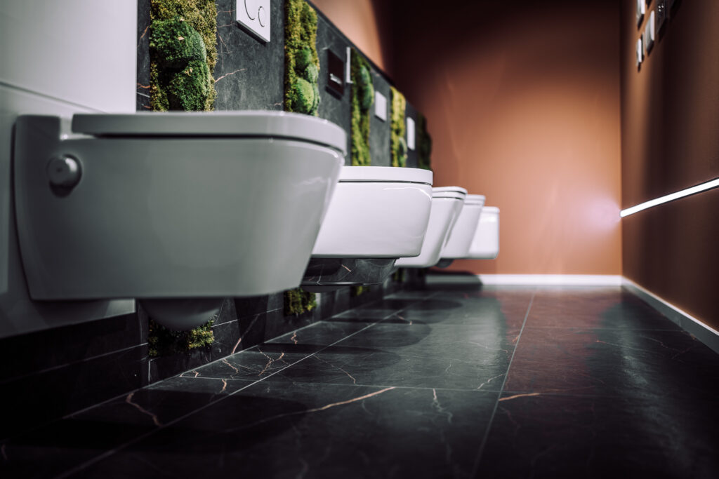 Verschiedene Toiletten mit unterschiedlichen Betätigungsplatten, an der Wand montiert, vor grauen Fliesen und dekorativem Moos.